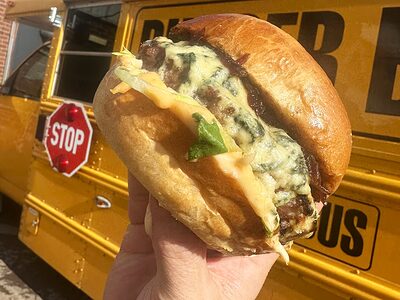 A cheeseburger held up in front of a yellow bus